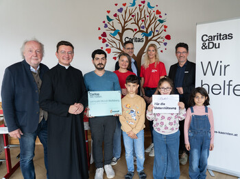 Gruppe aus Kindern, Freiwilligen und Unterstützer*innen im Lerncafé Mondi Wolfsberg vor Caritas-Rollup und Wandbild mit Herzen.