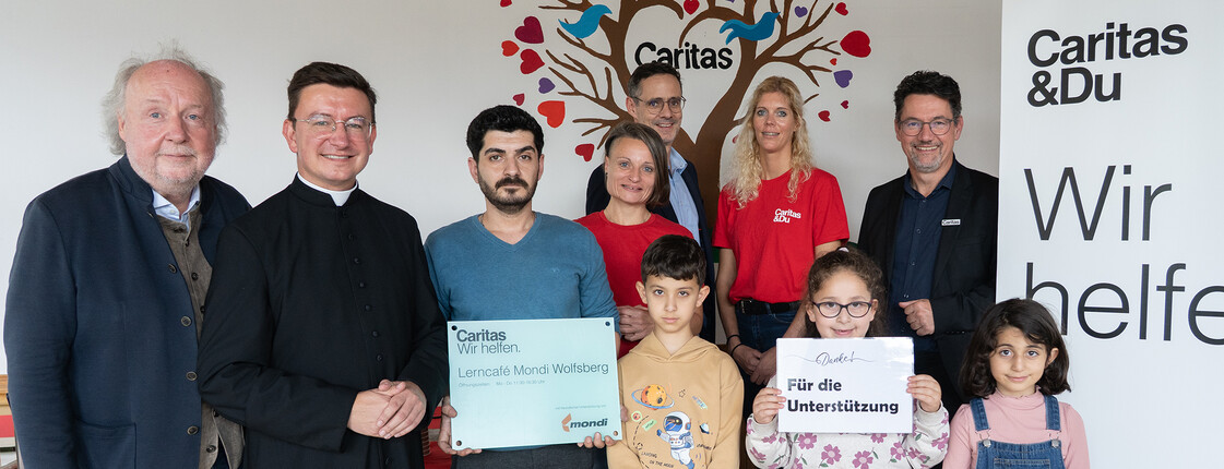 Gruppe aus Kindern, Freiwilligen und Unterstützer*innen im Lerncafé Mondi Wolfsberg vor Caritas-Rollup und Wandbild mit Herzen.