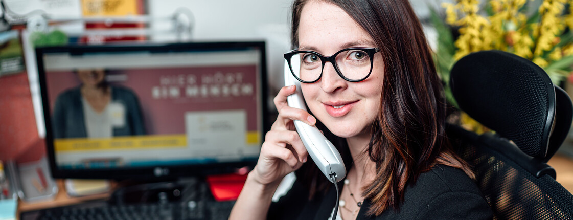 Eine junge Frau sitzt vor einem Computer und hält einen Telefonhörer in der Hand.
