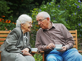 Zwei Menschen, ein Mann und eine Frau, die beide graue Haare haben, unterhalten sich gemütlich bei einer Tasse Kaffee im Garten. Der Mann trägt wie die Frau eine Brille. Er hat ein Hemd an und die Frau trägt einen grauen Mantel. 