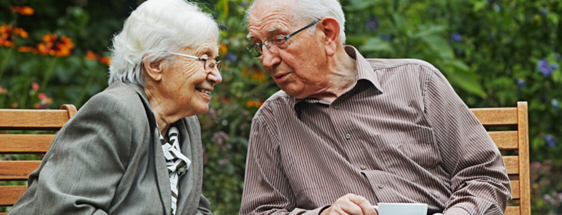 Zwei Menschen, ein Mann und eine Frau, die beide graue Haare haben, unterhalten sich gemütlich bei einer Tasse Kaffee im Garten. Der Mann trägt wie die Frau eine Brille. Er hat ein Hemd an und die Frau trägt einen grauen Mantel. 