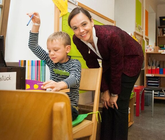 Ein Kind arbeitet mit Lernmaterialien an einem Tisch, während eine erwachsene Begleitperson den Unterricht im Klassenraum begleitet.