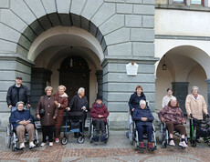 Menschen, einige in Rollstühlen, vor einem Gebäude mit gewölbten Türen und Steinfassade.