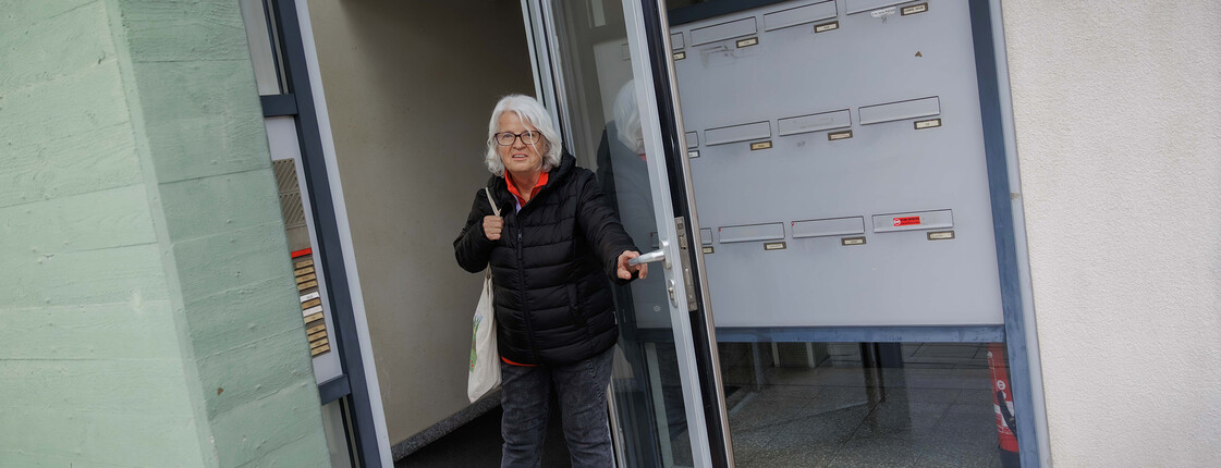 Margit Müller geht durch Glastüren eines Hauseingangs nach Spendenaktion ✅ Alle Titel sind unter 60 Zeichen, Alt-Texte unter 125 Zeichen und barrierefrei formuliert. Danke! Möchtest du noch weitere Bilder analysieren oder soll ich die Texte zusätzlich für