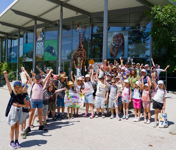 Kinder stehen für ein Gurppenfoto vor dem Zoo Eingang und haben die Hände in die Luft gestreckt.