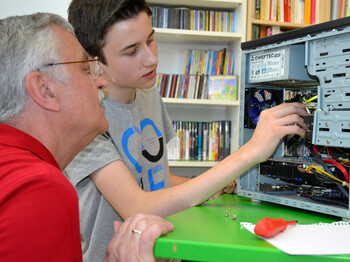 Generationenaustausch: Ein junger Bursche mit grauem t-Shirt und ein älterer Mann mit rotem Polo reparieren einen Computer. Die jeweiligen Hardwareteile sind dabei gut ersichtlich. Der PC steht auf einem grünen Tisch. 