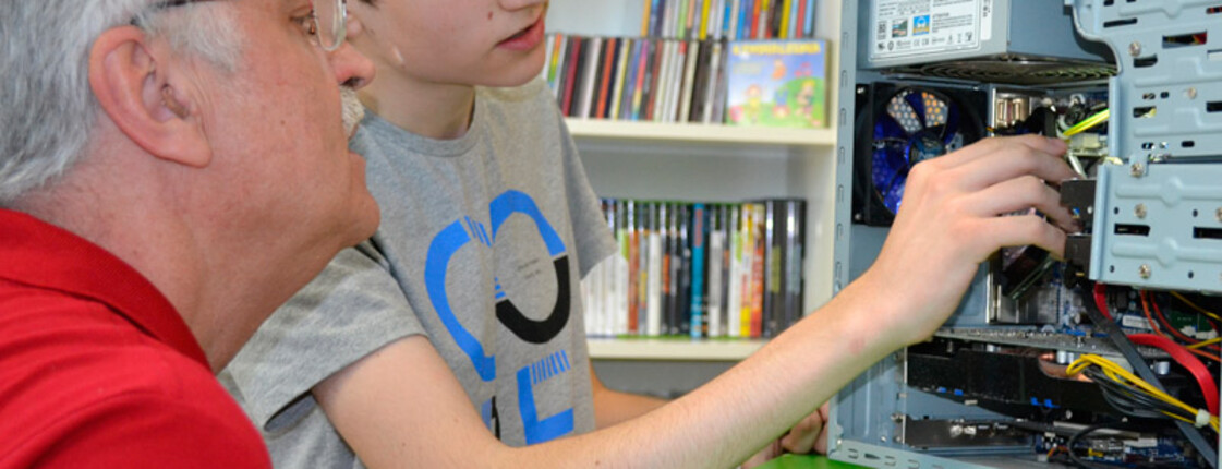 Generationenaustausch: Ein junger Bursche mit grauem t-Shirt und ein älterer Mann mit rotem Polo reparieren einen Computer. Die jeweiligen Hardwareteile sind dabei gut ersichtlich. Der PC steht auf einem grünen Tisch. 