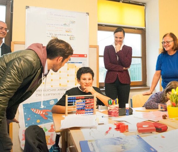 Kinder arbeiten gemeinsam im Klassenraum mit Lernmaterialien, während mehrere Erwachsene die Unterrichtssituation begleiten.