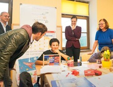 Kinder arbeiten gemeinsam im Klassenraum mit Lernmaterialien, während mehrere Erwachsene die Unterrichtssituation begleiten.
