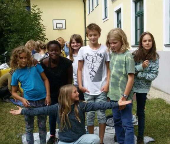 Kinder stehen bei einer Teamaufgabe gemeinsam auf einer Plane im Schulgarten und fördern spielerisch Zusammenarbeit sowie Geschicklichkeit.