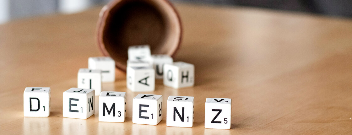 Eine brauner Becher liegt umgekippt auf einem Holztisch. Daneben liegen Würfel mit Buchstaben. Sie bilden das Wort „DEMENZ“.