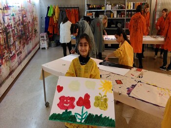 Mehrere Kinder und Jugendliche gestalten Bilder in einem Atelier. Im Vordergrund hält eine Person ein selbst gemaltes Bild, im Hintergrund wird an weiteren Kunstwerken gearbeitet.