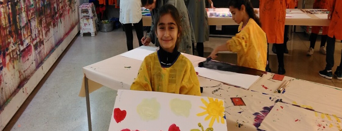 Mehrere Kinder und Jugendliche gestalten Bilder in einem Atelier. Im Vordergrund hält eine Person ein selbst gemaltes Bild, im Hintergrund wird an weiteren Kunstwerken gearbeitet.