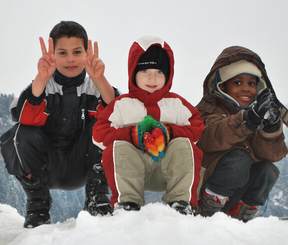 Drei Kinder unterschiedlicher Ethnien spielen im Schnee. Sie tragen dicke Winterbekleidung, die schwarz, weiß, rot und braun ist. Sie sitzen dabei in einer Hocke auf einem Schneehügel. Alle drei Kinder sind erfreut und haben Spaß. Einer der drei Jungs zeigt noch das Victory Zeichen mit beiden Händen. 