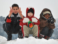 Drei Kinder unterschiedlicher Ethnien spielen im Schnee. Sie tragen dicke Winterbekleidung, die schwarz, weiß, rot und braun ist. Sie sitzen dabei in einer Hocke auf einem Schneehügel. Alle drei Kinder sind erfreut und haben Spaß. Einer der drei Jungs zeigt noch das Victory Zeichen mit beiden Händen. 