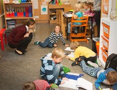 Kinder arbeiten mit Arbeitsblättern auf dem Boden und an Tischen, während eine Lehrkraft die gemeinsame Lernaktivität begleitet.