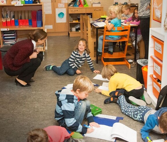 Kinder arbeiten mit Arbeitsblättern auf dem Boden und an Tischen, während eine Lehrkraft die gemeinsame Lernaktivität begleitet.