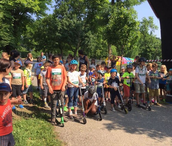 Kinder stehen mit Rollern am Start einer Sportveranstaltung im Freien und bereiten sich gemeinsam auf einen Wettbewerb vor.