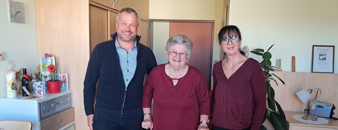Bürgermeister Herr Tulnik und Pflegedienstleitung Frau Simko stehen gemeinsam mit Frau Krammer in ihrem Zimmer. Alle drei lächeln in die Kamera anlässlich ihres 90. Geburtstags.