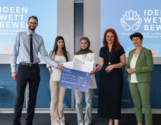 Fünf Personen stehen vor zwei Leinwänden mit Projektionen auf einer Bühne. Zwei Schülerinnen in der Mitte halten Urkunden hoch.