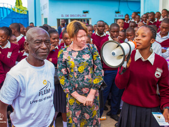Eine große Gruppe von Schülerinnen und Schülern steht im Schulhof. Im Vordergrund spricht ein Mädchen in Schuluniform durch ein Megafon. Neben ihr stehen ein älterer Mann im FLEM-T-Shirt und eine Frau in einem geblümten Kleid. Alle blicken aufmerksam in Richtung der Sprecherin.