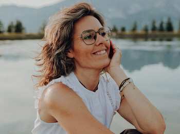 Hochformatiges Portrait einer Frau mittleren Alters, die vor einem Bergsee sitzt und ihren Kopf auf dem linken Arm abstützt.