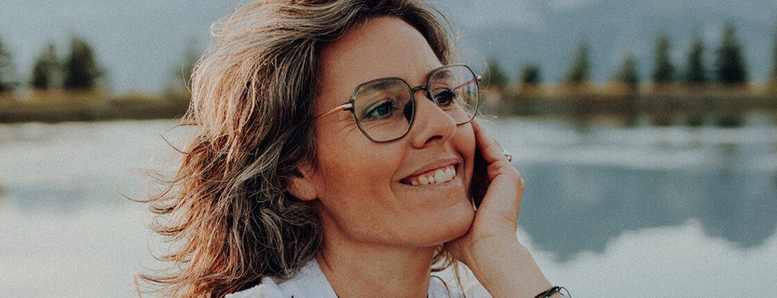 Hochformatiges Portrait einer Frau mittleren Alters, die vor einem Bergsee sitzt und ihren Kopf auf dem linken Arm abstützt.