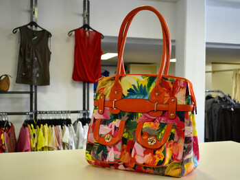 Eine rote Handtasche mit Blumenmuster steht auf einem hölzernen Tisch. Im Hintergrund sind Kleider an Kleiderhaken. 
