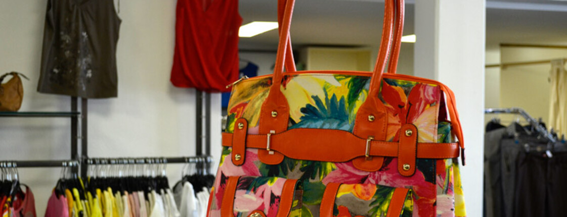 Eine rote Handtasche mit Blumenmuster steht auf einem hölzernen Tisch. Im Hintergrund sind Kleider an Kleiderhaken. 