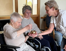 Seniorin in Rollstuhl schlägt Klangschale, unterstützt von einer Yoga-Anleiterin, zur Meditation.