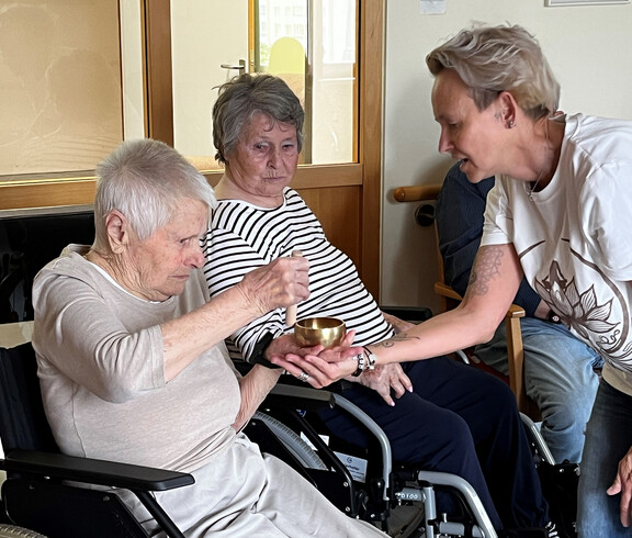 Seniorin in Rollstuhl schlägt Klangschale, unterstützt von einer Yoga-Anleiterin, zur Meditation.