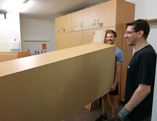 Zwei Personen tragen ein großes Möbelteil durch einen Innenraum und unterstützen gemeinsam bei Umbau- oder Einrichtungsarbeiten.