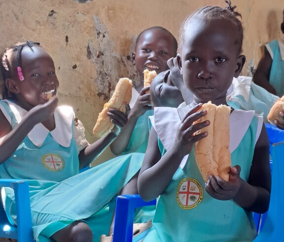 Drei südsudanesische Mädels blicken in die Kamera. Sie tragen Schuluniform und essen eine Schuljause. 