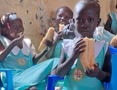 Drei südsudanesische Mädels blicken in die Kamera. Sie tragen Schuluniform und essen eine Schuljause. 
