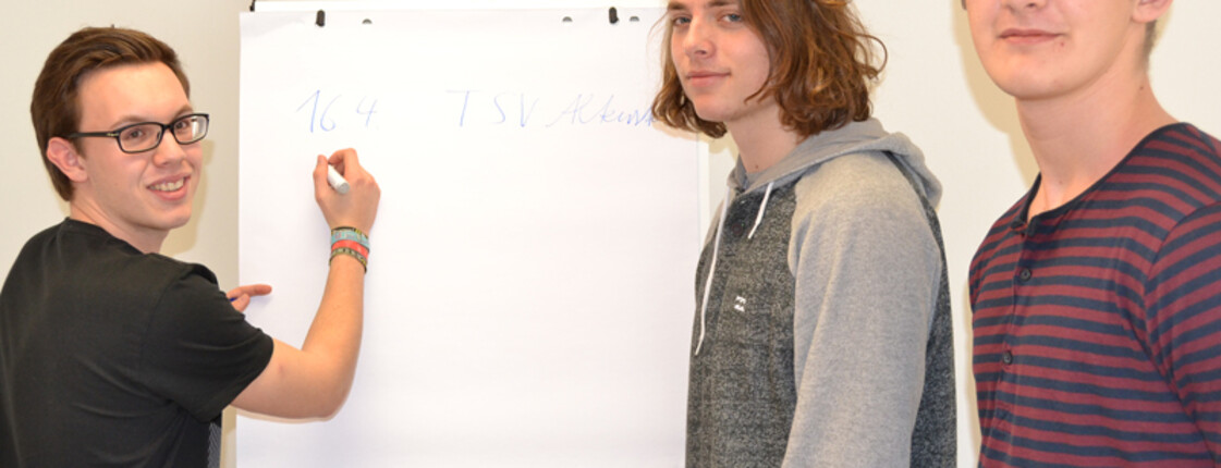 Drei junge Erwachsene stehen vor einem Flipchart. Einer davon schreibt etwas auf eine Seite des Flipchartes. Die anderen beiden schauen in die Kamera.