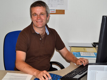 Ein Herr mittleren Alters sitzt vor einem Computer. Sein Schreibtisch ist sehr aufgeräumt. 