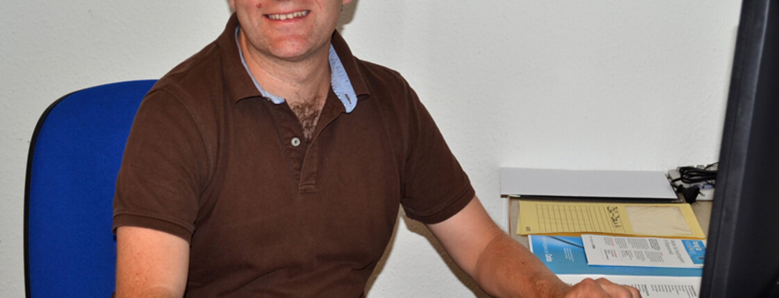 Ein Herr mittleren Alters sitzt vor einem Computer. Sein Schreibtisch ist sehr aufgeräumt. 