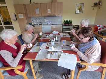Bewohnerinnen des Pflegewohnhauses bemalen gemeinsam Keramikfiguren und gestalten farbenfrohe Osterdekorationen an einem Arbeitstisch.