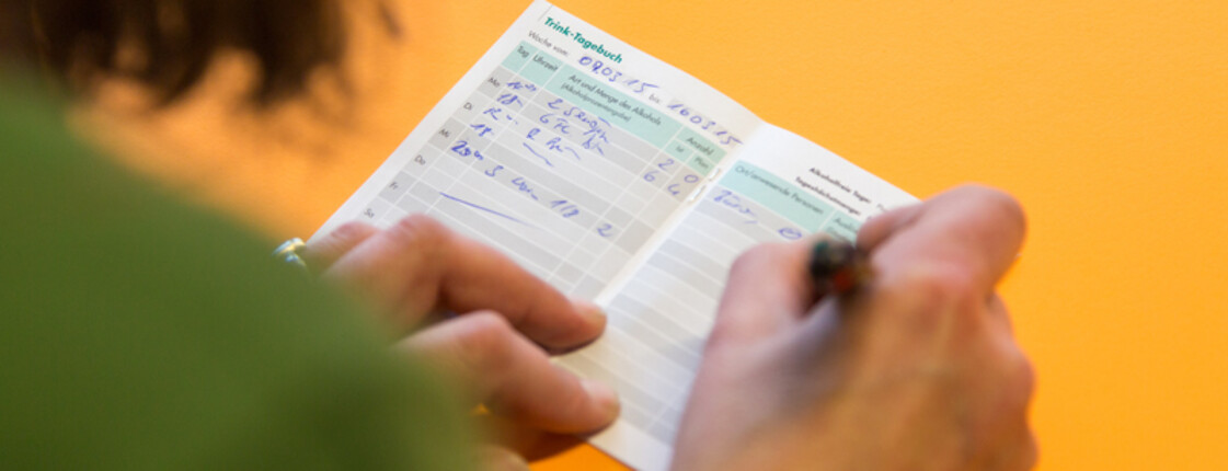 Eine Person mit grünem Oberteil notiert etwas in einem kleinen Heftchen mit der Aufschrift "Trink-Tagebuch".