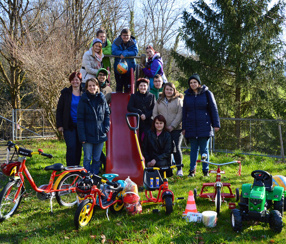 Eine Gruppe junger Erwachsener steht auf einem Kinderspielplatz und zeigt stolz ihre Sachspenden für MUKI. Darunter sind unter anderem Fahrräder und Spielzeugtraktoren. 