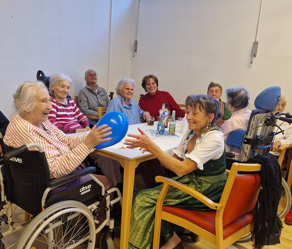 Senior*innen am Tisch, eine Frau im Rollstuhl spielt lachend mit einem Ballon, festliche Stimmung