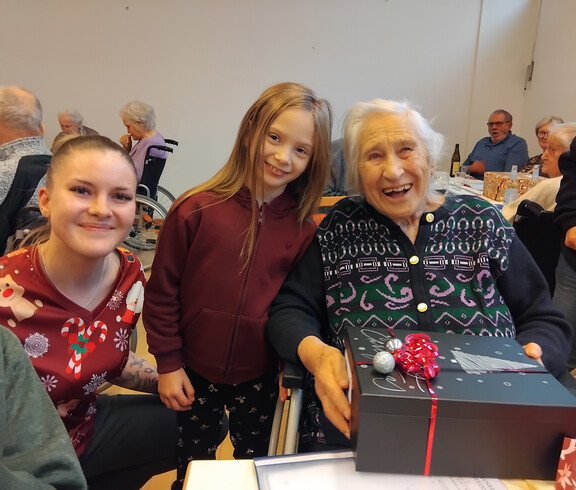 Bewohnerin hält ein schwarzes Geschenk mit roter Schleife, daneben eine junge Dame und en Mädchen.
