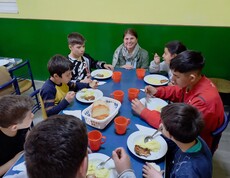 Eine Gruppe Kinder sitzt am Tisch und isst. Eine Frau kniet daneben. 