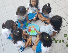 Sechs kleine Mädchen in blau-weißer Schuluniform sitzen im Kreis auf dem Boden und spielen gemeinsam mit bunten Spielzeugfischen in einer Schüssel mit Wasser.