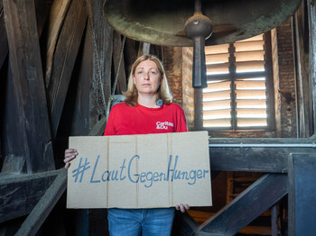 Caritas-Präsidentin Nora Tödtling-Musenbichler hält ein Schild mit dem #LautGegenHunger. Sie steht vor einer Glocke in einem Kirchturm.