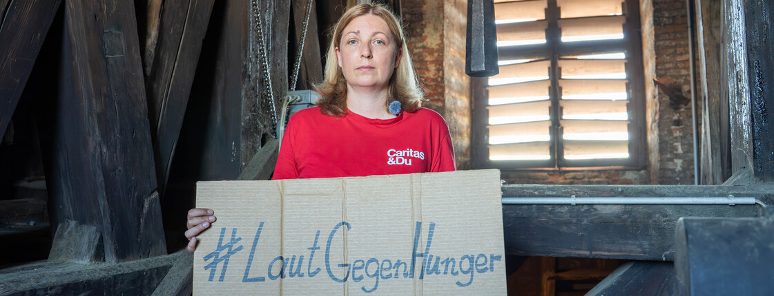 Caritas-Präsidentin Nora Tödtling-Musenbichler hält ein Schild mit dem #LautGegenHunger. Sie steht vor einer Glocke in einem Kirchturm.
