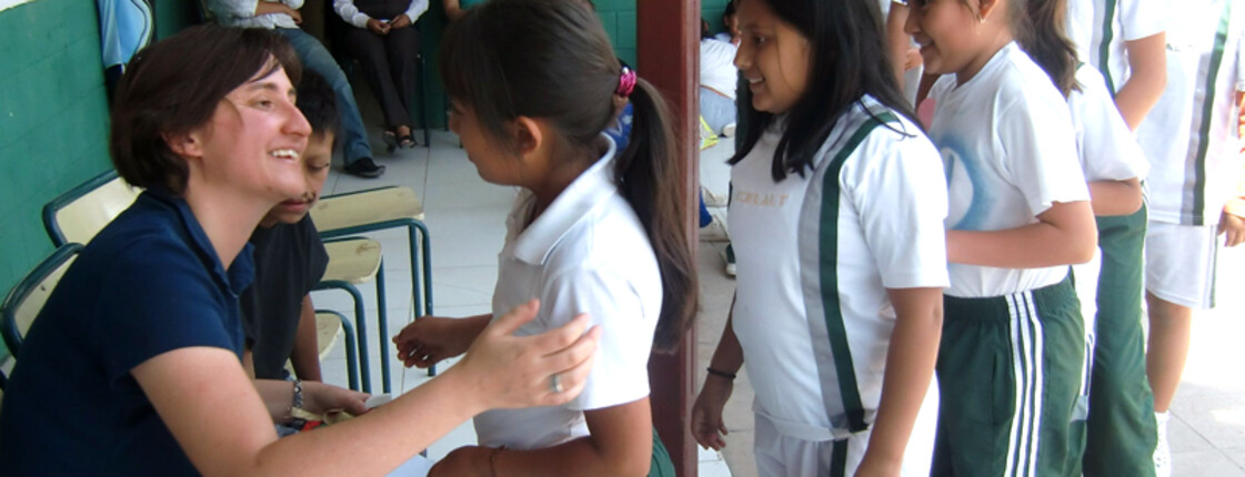 Eine Frau sitzt auf einem Stuhl und begrüßt herzlich die Schülerinnen von Ecuador. Die Kinder tragen alle eine Schuluniform und stehen in einer Reihe.