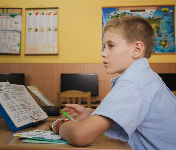 Ein kleiner junge sitzt an seinem Tisch in einem Klassenzimmer. Vor ihm steht ein offenes Lesebuch.