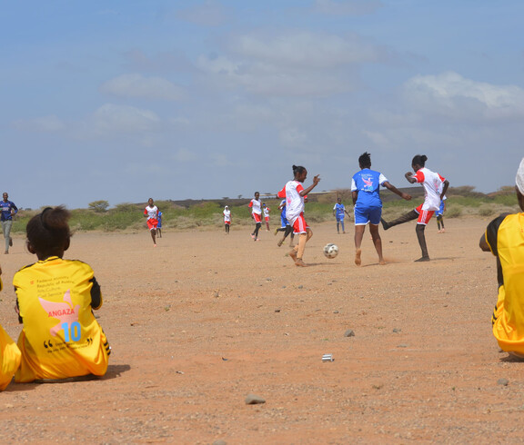 Mehrere Afrikaner*innen spielen auf einem sandigen Platz Fußball. Zwei Teams in unterschiedlichen Trikots treten gegeneinander an, während eine weitere Person am Spielfeldrand sitzt und zuschaut.
