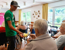 Jugendlicher verteilt Eis an mehrere Seniorinnen im Rollstuhl im Aufenthaltsraum